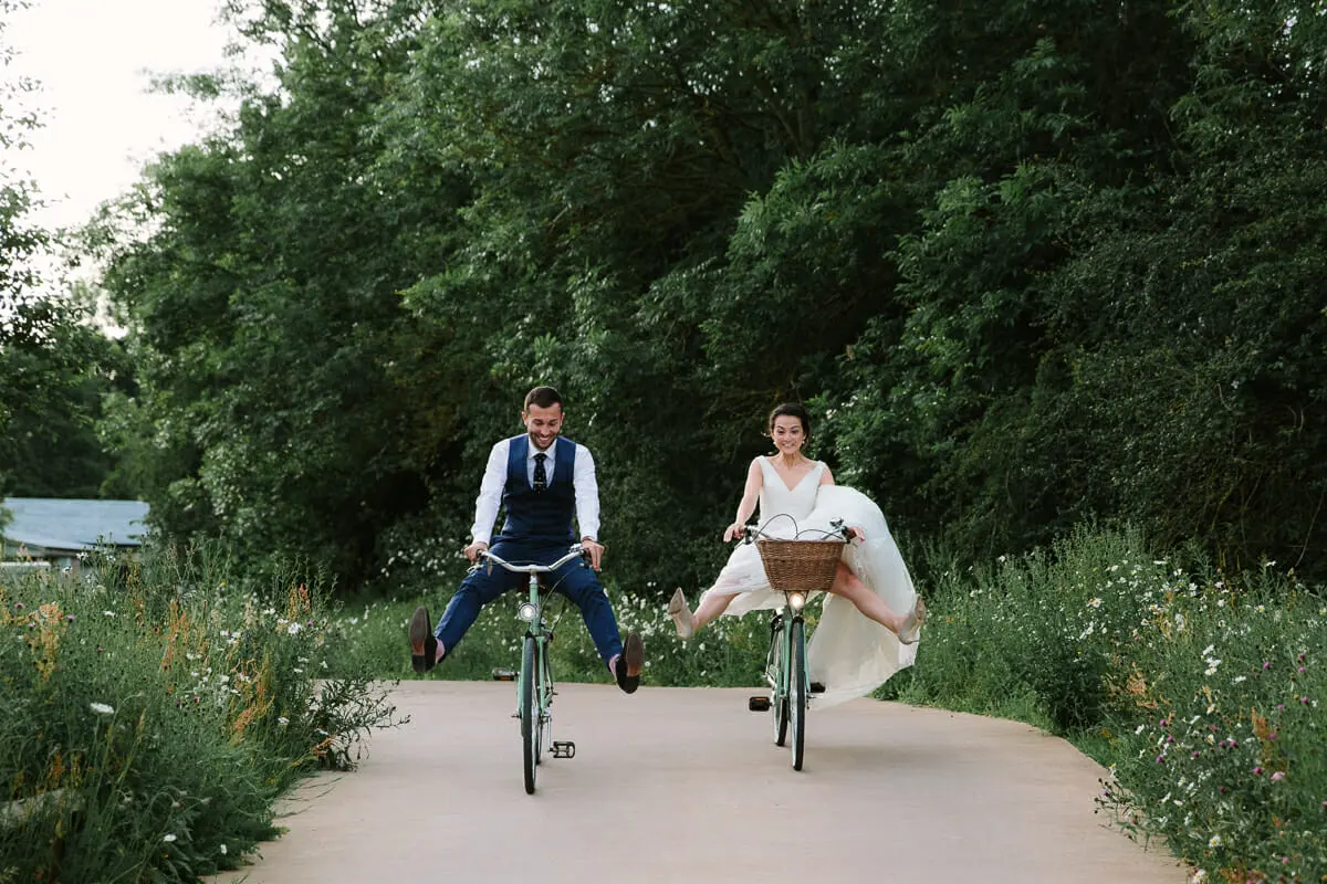 Bride and groom riding bicycles with their legs out along a path at Soho Farmhouse