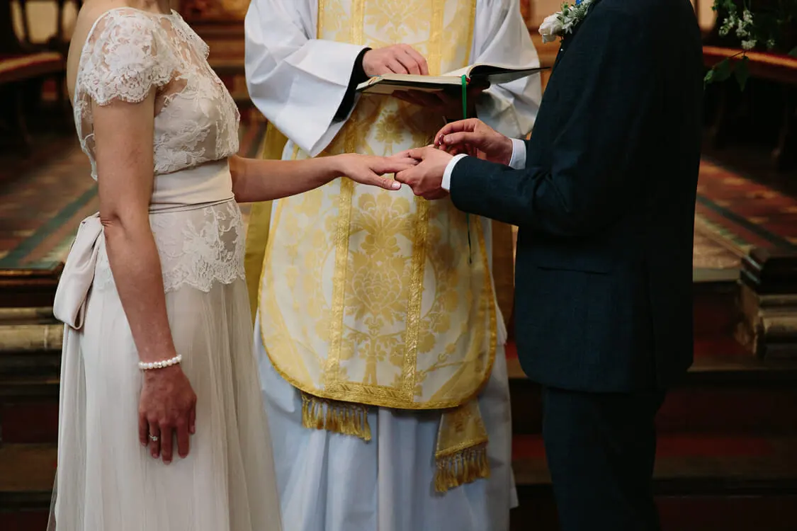 Groom placing a ring on the bride's finger in front of a priest in gold vestments