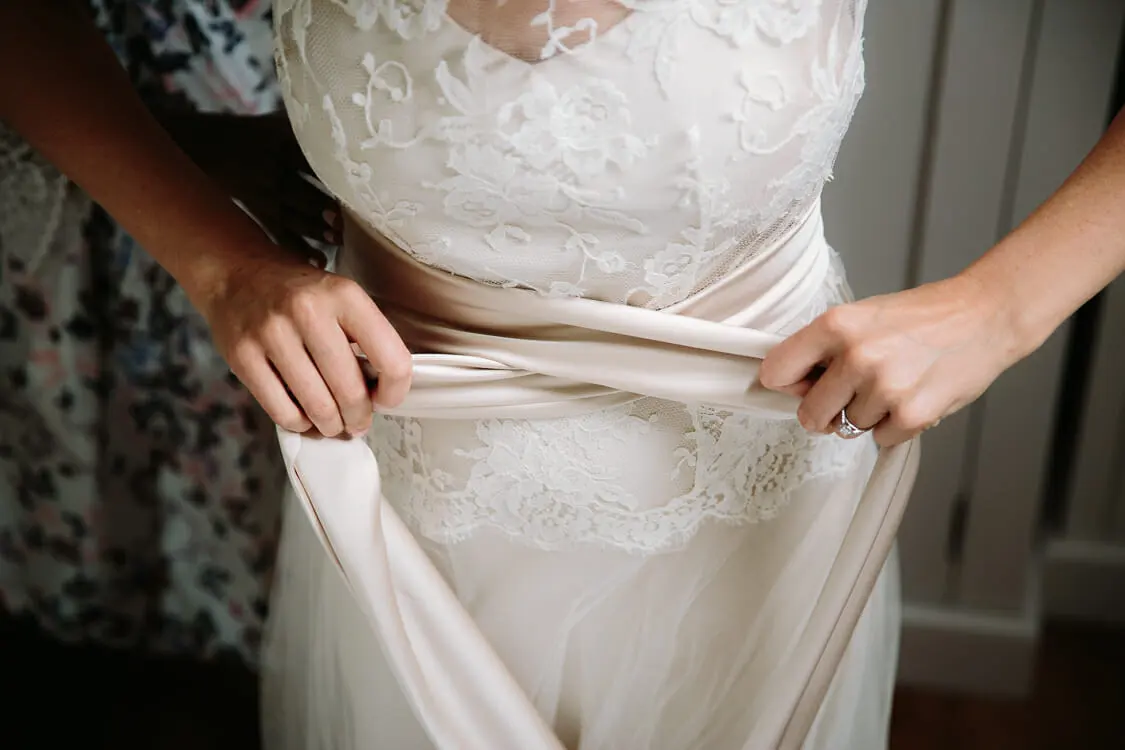 Hands tying a blush satin sash around a bride's lace wedding dress while she gets ready