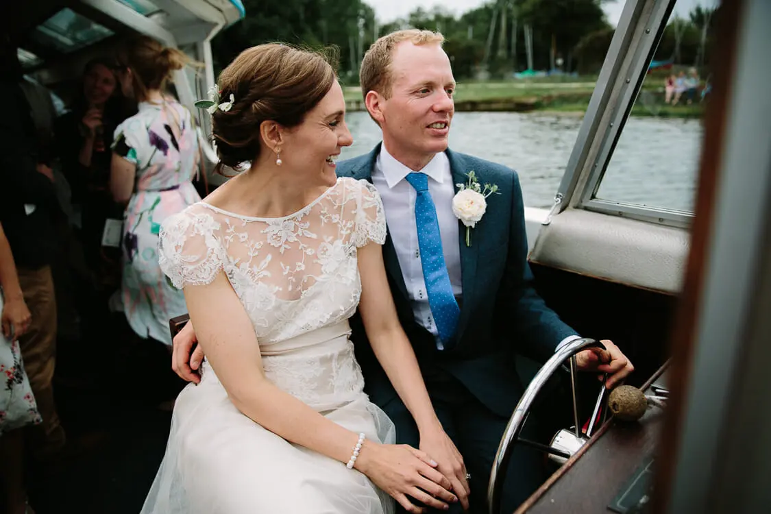 Bride and groom smiling at the wheel of a river boat as guests chat behind them