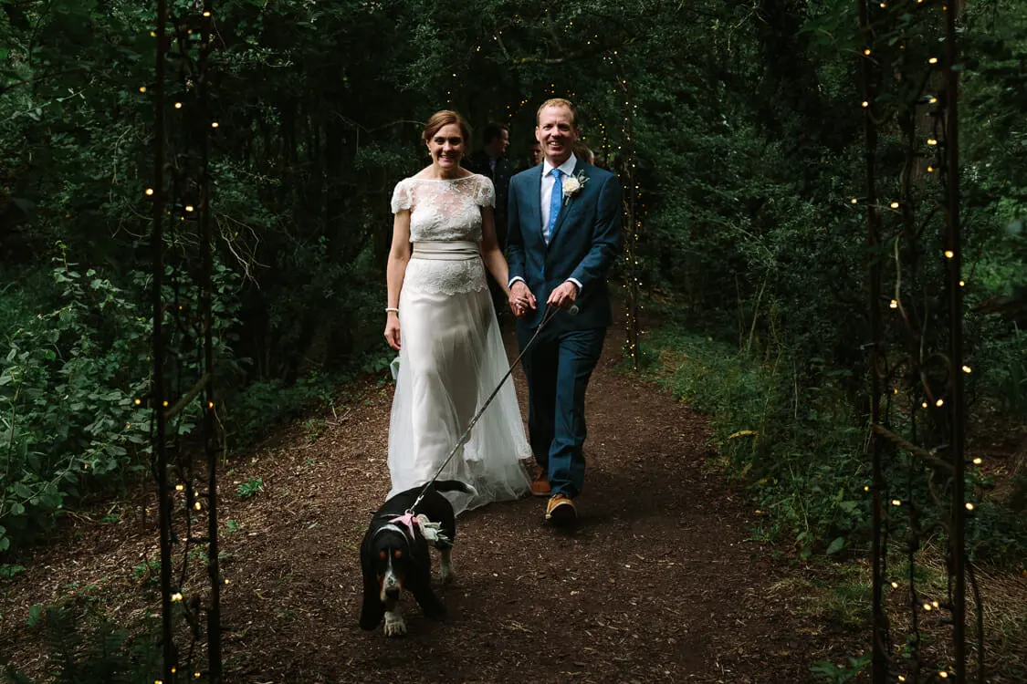 Bride and groom walking a basset hound down a woodland path strung with fairy lights at the Perch