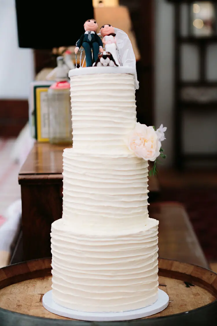 Three-tier white buttercream cake with a peach rose and bride, groom and dog toppers on a barrel