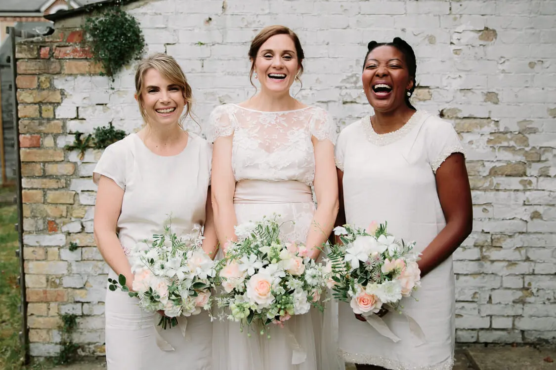 Bride and two bridesmaids in cream laughing with blush and white bouquets against a whitewashed brick wall