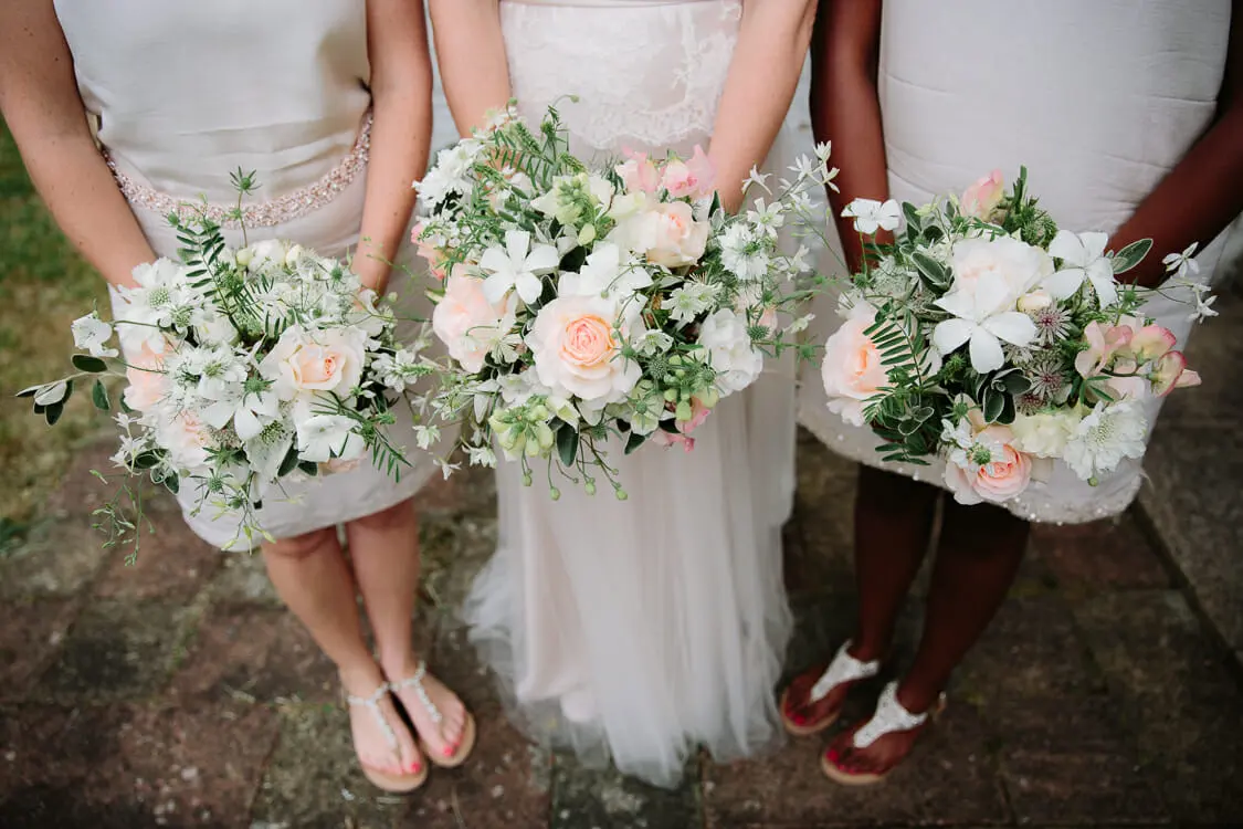 Bride and two bridesmaids holding white and peach bouquets of roses and greenery, seen from the waist down
