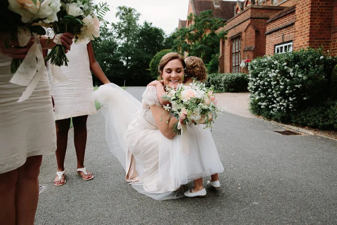 Bride crouching to hug a young flower girl on a driveway outside a red brick building