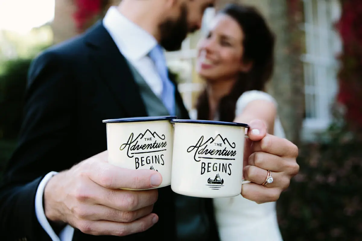 Couple clinking enamel mugs printed with The Adventure Begins, her engagement ring in view