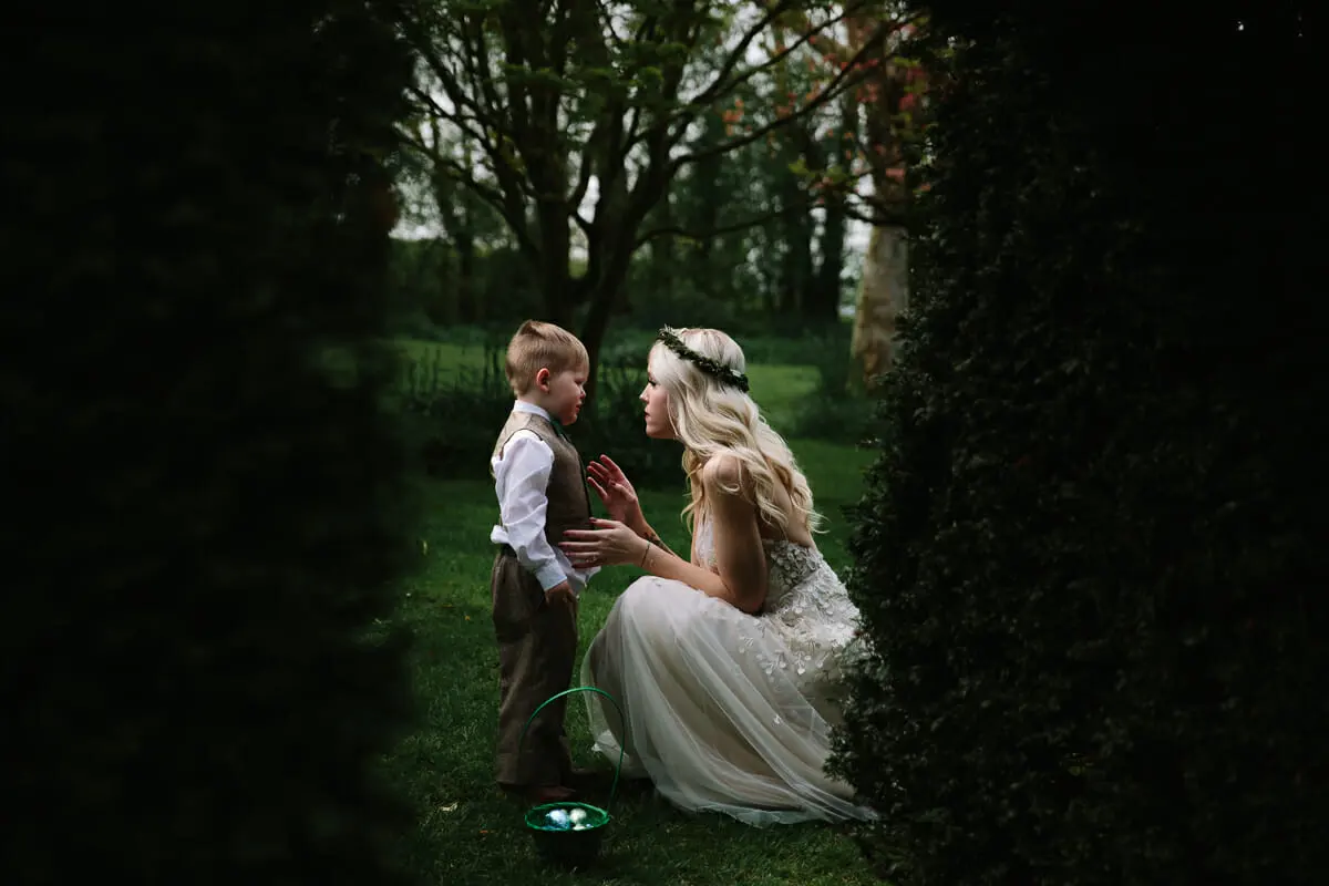 Bride in a flower crown kneeling on the lawn to talk with a little page boy between dark hedges