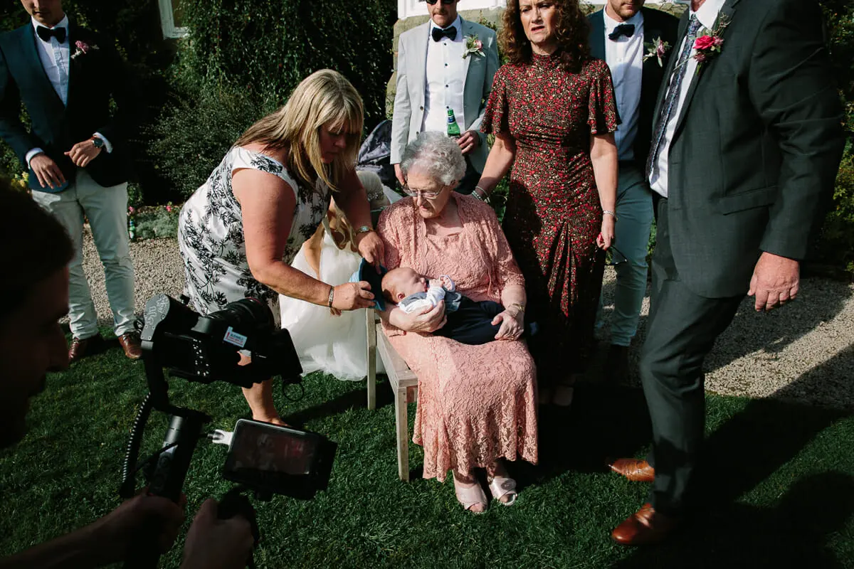 Grandmother holding a baby on a garden chair as family gather round for a group photograph
