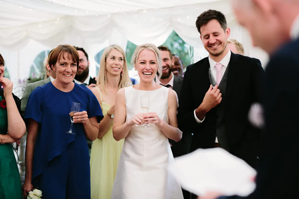 Bride with a glass of fizz laughing alongside the groom and guests during speeches in a marquee