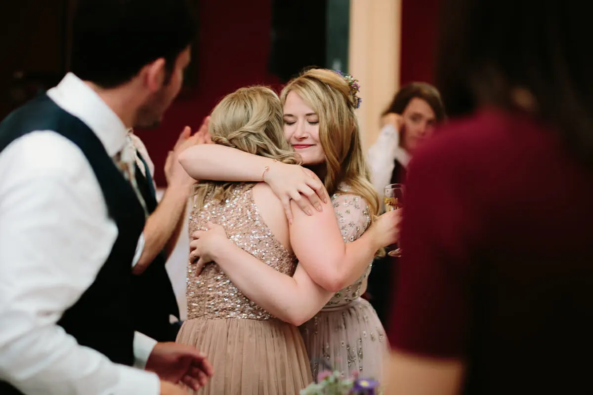 Two bridesmaids in sequinned dresses hugging during the reception as the groom looks on
