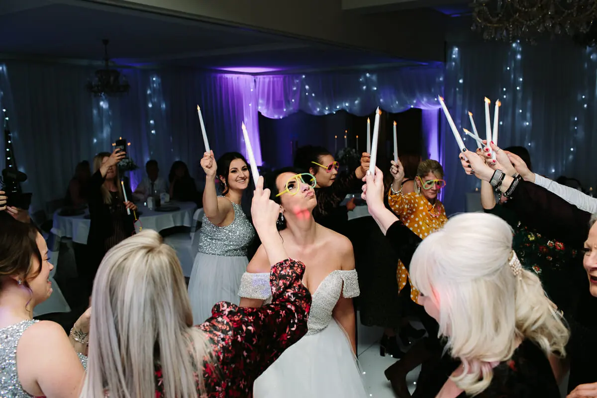 Bride in novelty glasses dancing with friends holding up candles on a lit dance floor