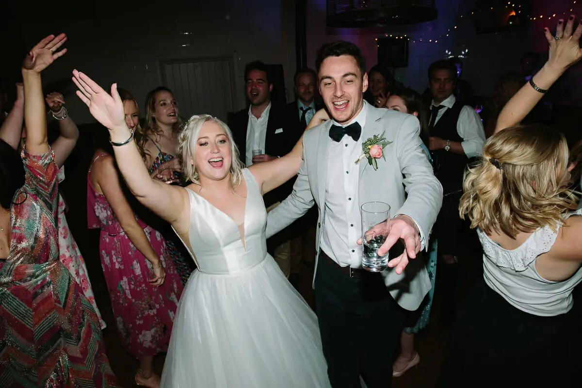 Bride and groom dancing with guests, arms in the air, on a crowded evening dance floor
