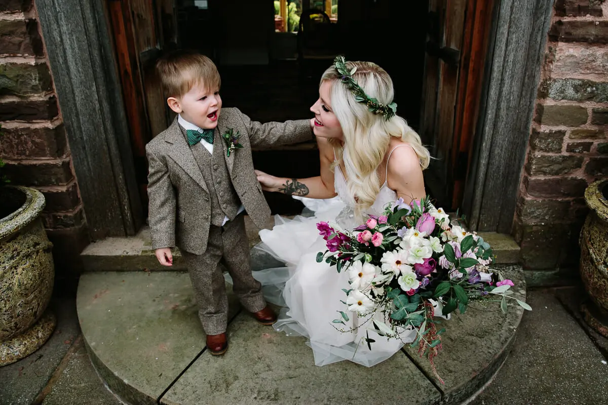 Bride in a leafy crown laughing with a page boy in a tweed suit on a doorstep