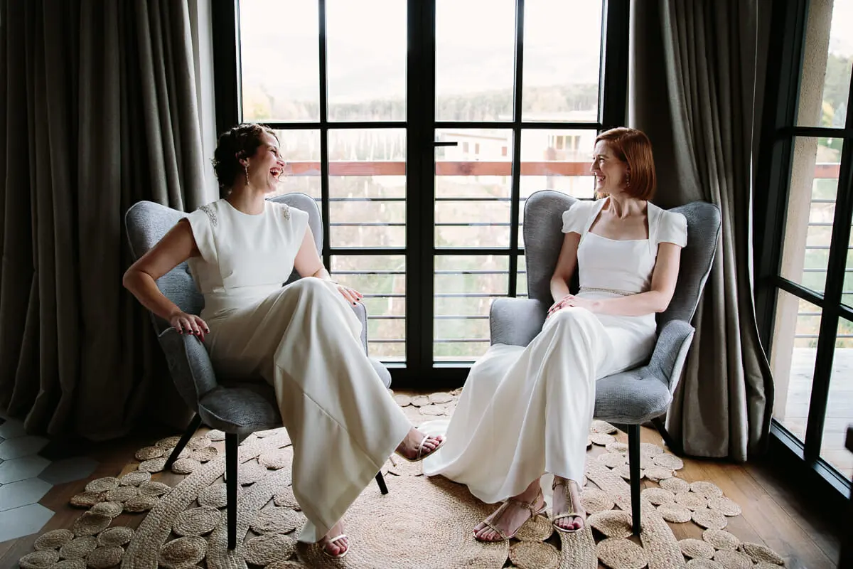 Two brides in white laughing together in armchairs by tall windows