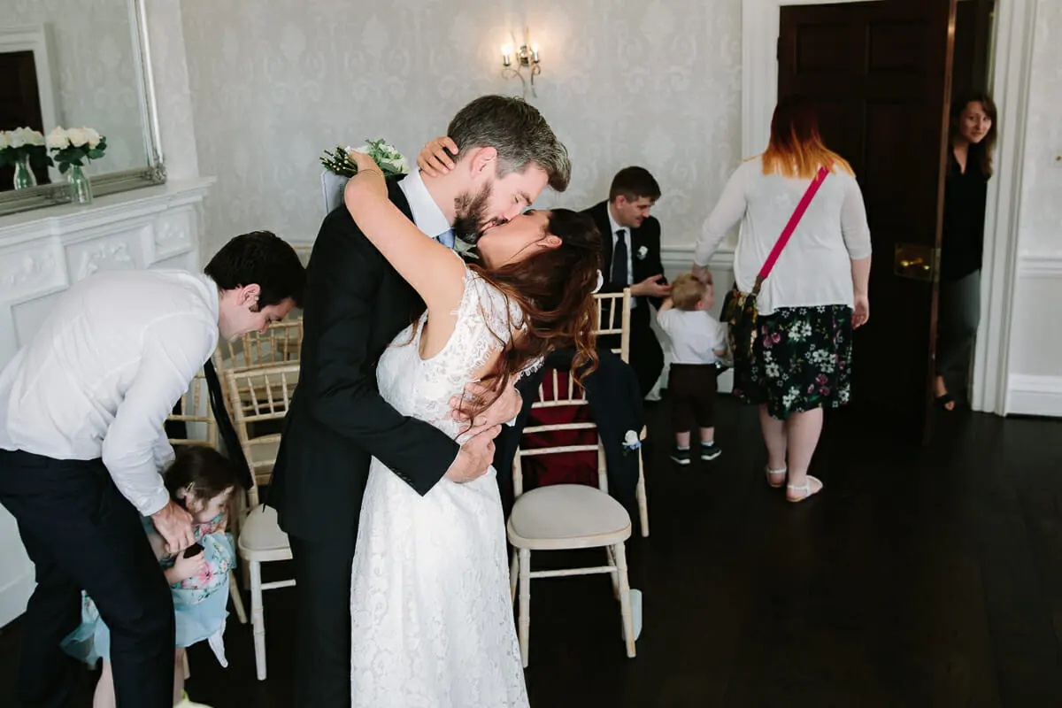 Bride and groom kissing among white chairs in an elegant room as guests leave after the ceremony