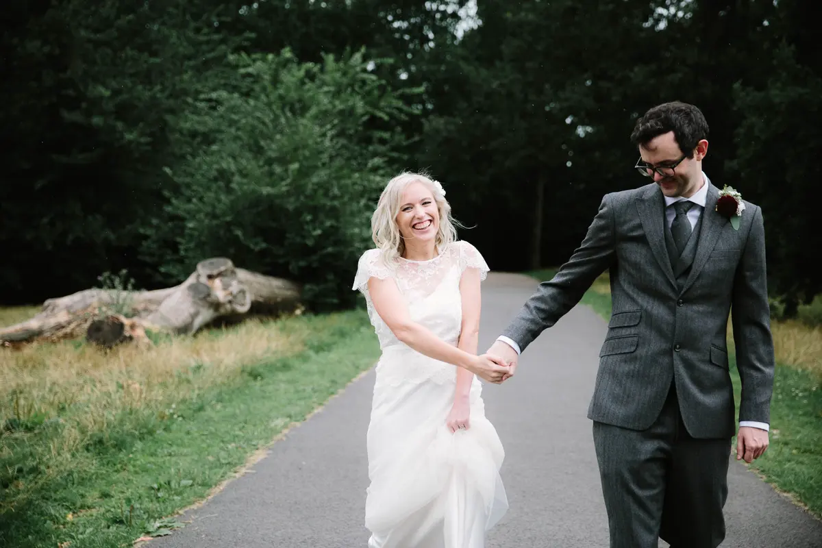 Bride laughing as she holds the groom's hand on a tree-lined path