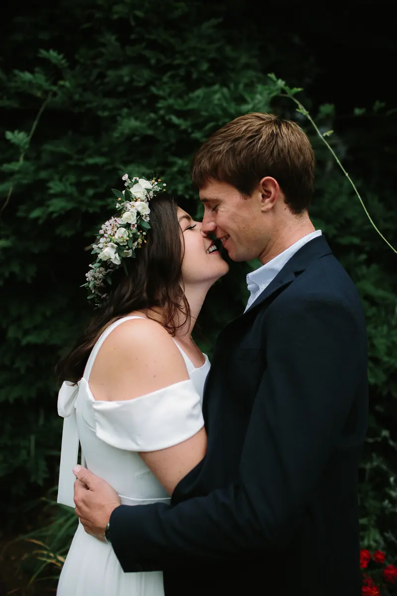Bride in a flower crown and groom in a navy suit nuzzling noses in front of dark greenery