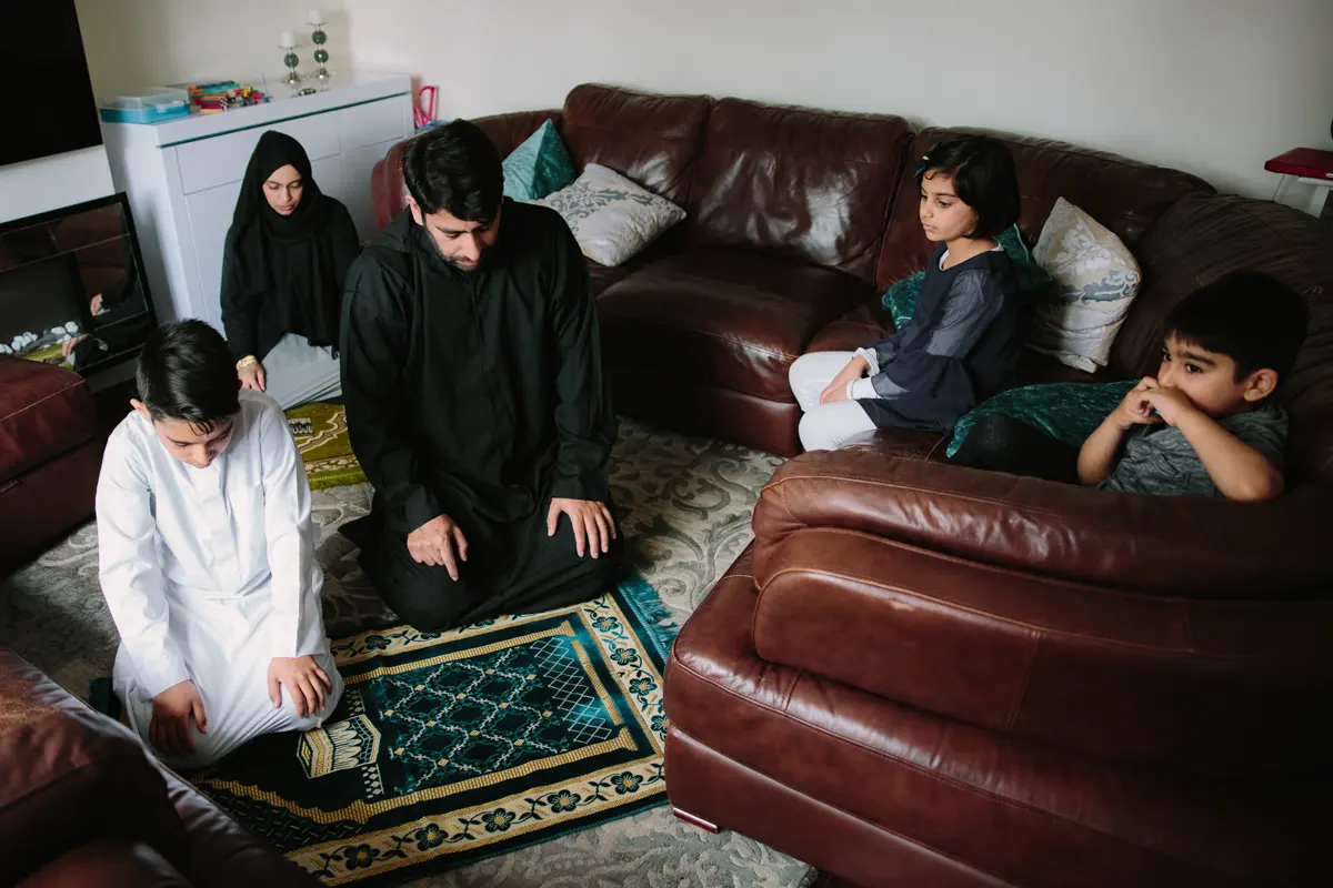 Father and son praying on prayer mats in a living room while family members sit nearby