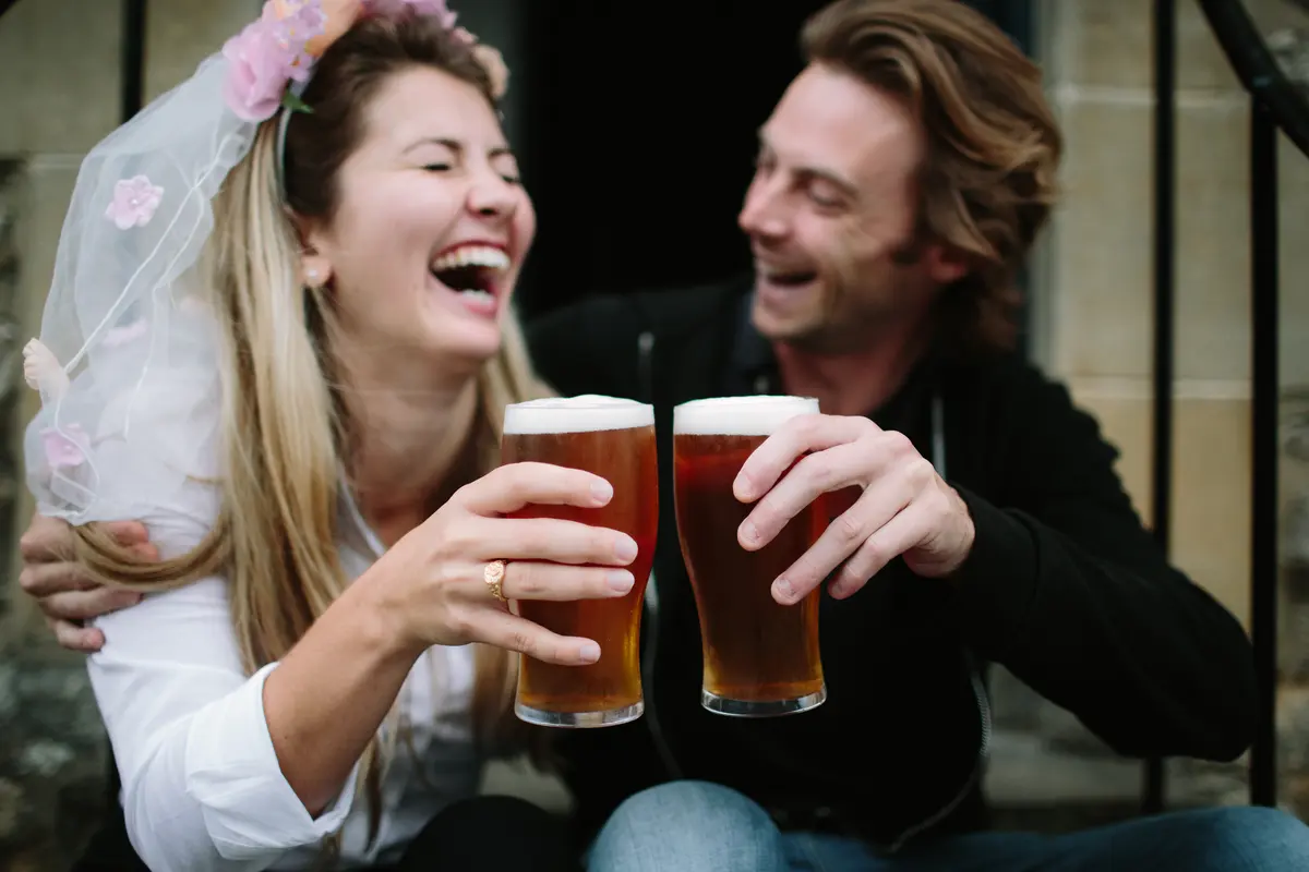 Laughing couple on stone steps clinking pints of beer, the woman wearing a short veil with pink flowers