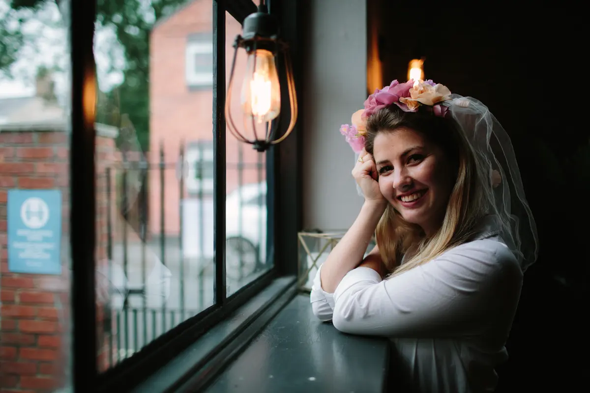 Smiling woman in a flower crown and short veil leaning on a window ledge in a pub
