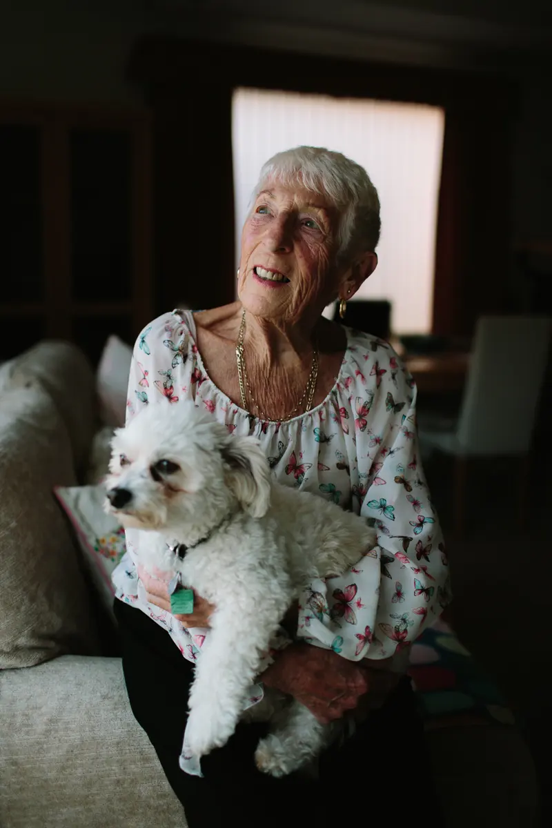 Older woman smiling as she sits on a sofa holding her small white dog