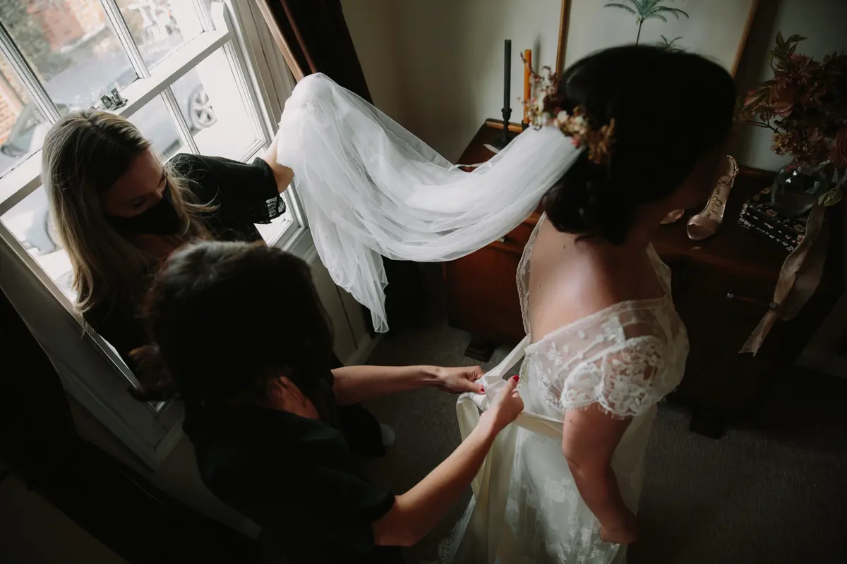 Two women in face masks helping a bride into her lace dress as her long veil is lifted