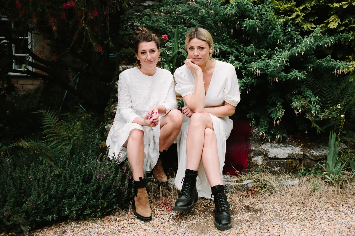 Two women in white dresses and boots sitting together on a garden wall
