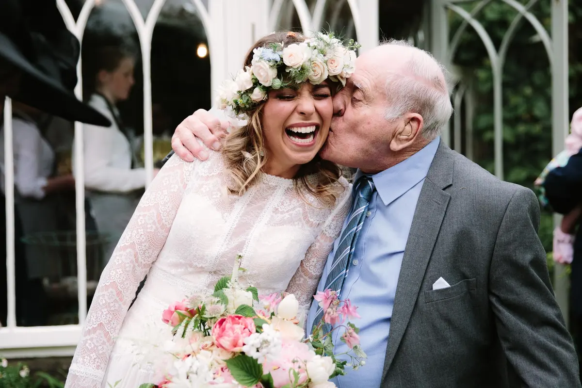 Older man kissing the laughing bride on the cheek as she holds her bouquet