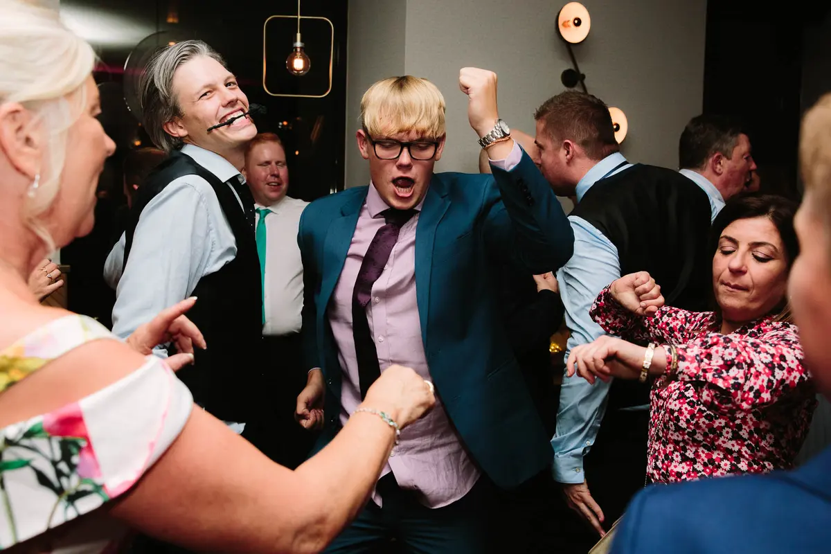 Guest in glasses and a teal suit punching the air on a crowded dance floor