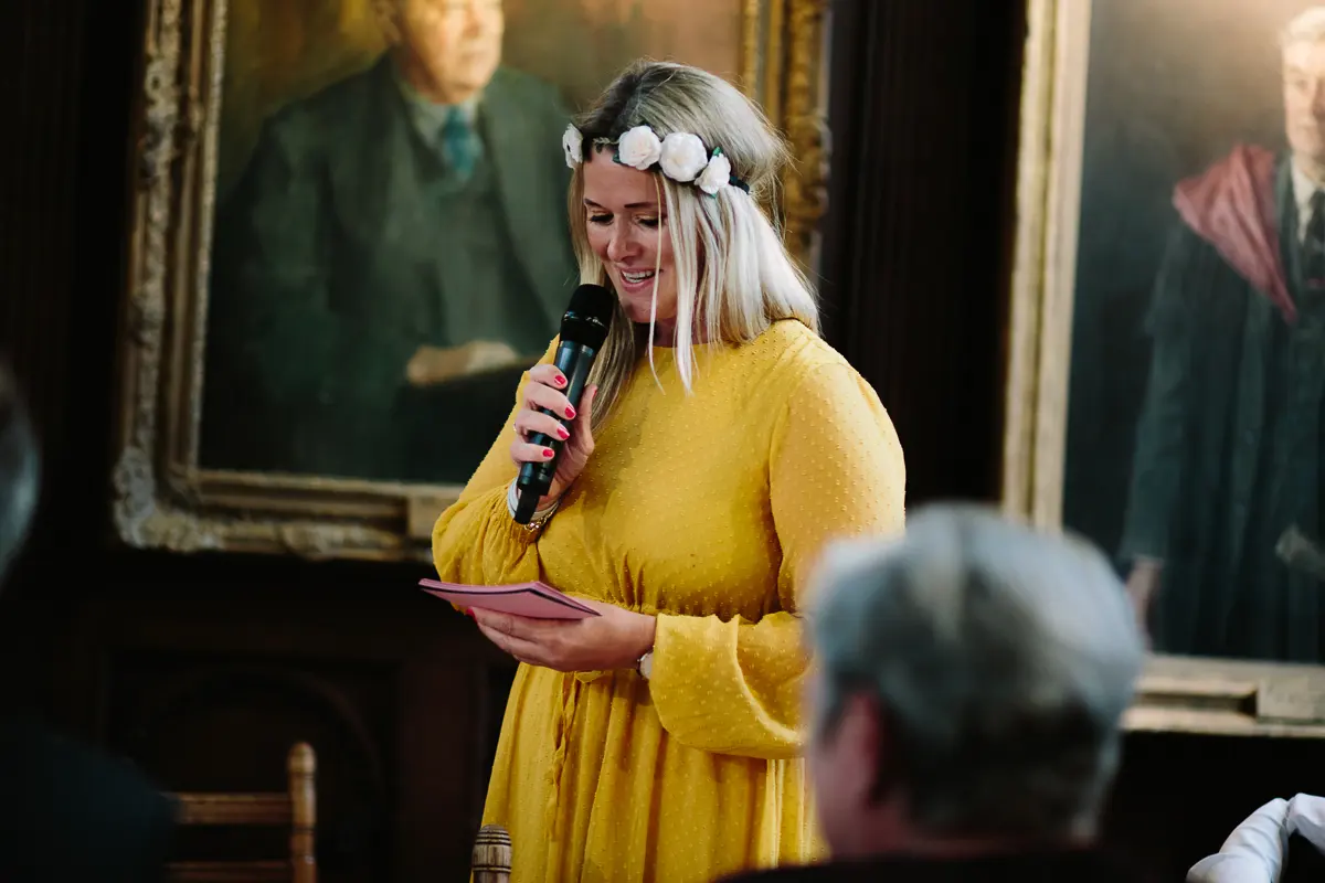 Woman in a yellow dress and flower crown reading a speech beneath oil portraits