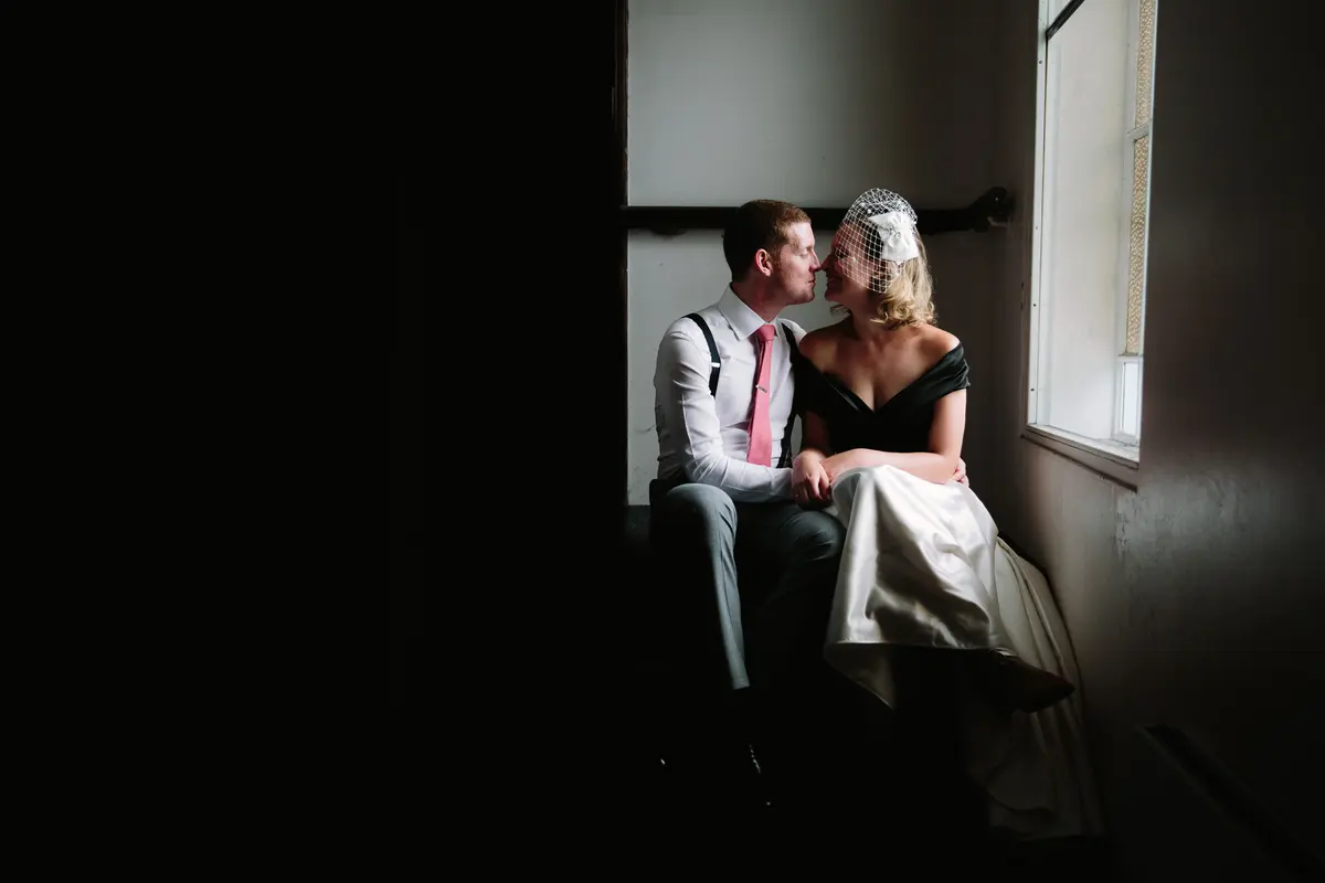 Bride in a birdcage veil and groom about to kiss on a window seat in soft light