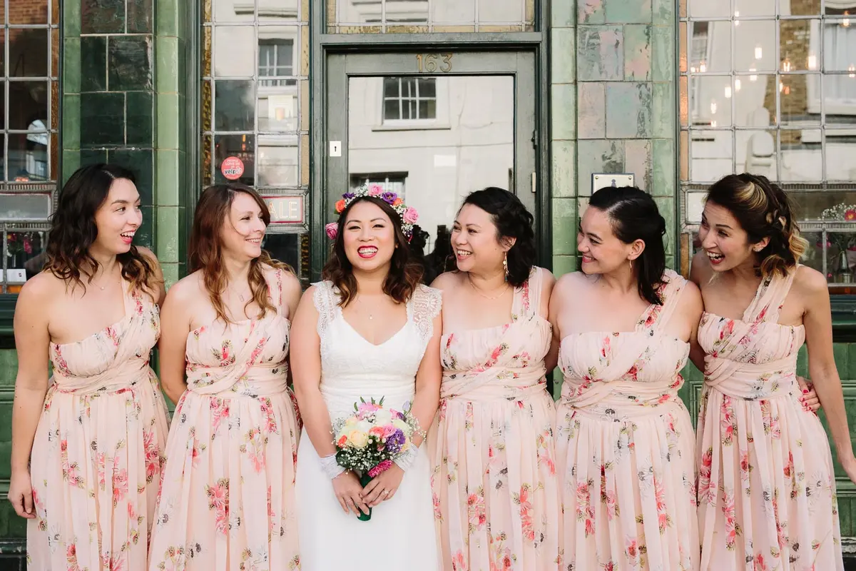 Bride in a flower crown with five bridesmaids in floral dresses outside a green tiled pub