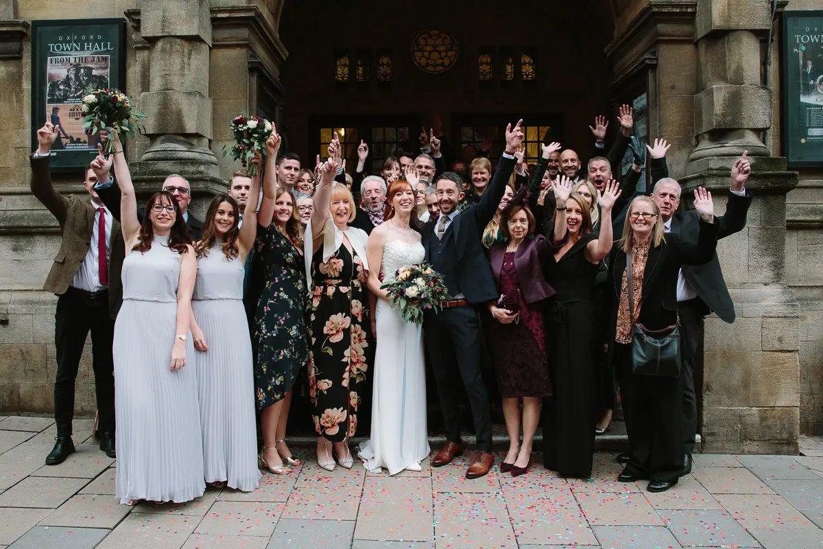 Wedding party cheering with arms raised on the steps of Oxford Town Hall amid confetti