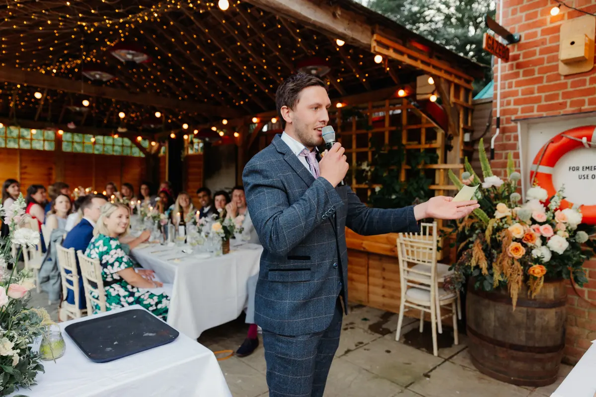 Best man giving a speech with a microphone under fairy lights at Cherwell Boathouse