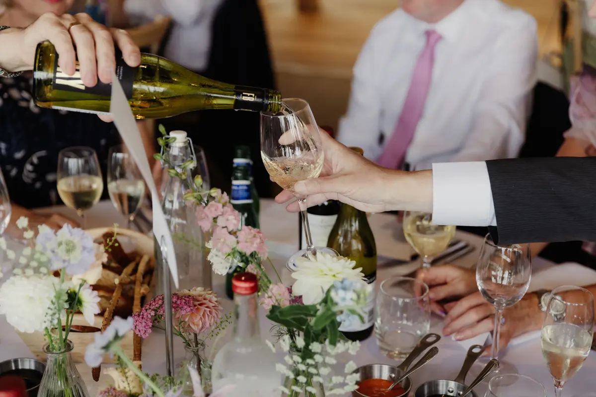 Guest pouring white wine into a glass held across the table at the wedding breakfast