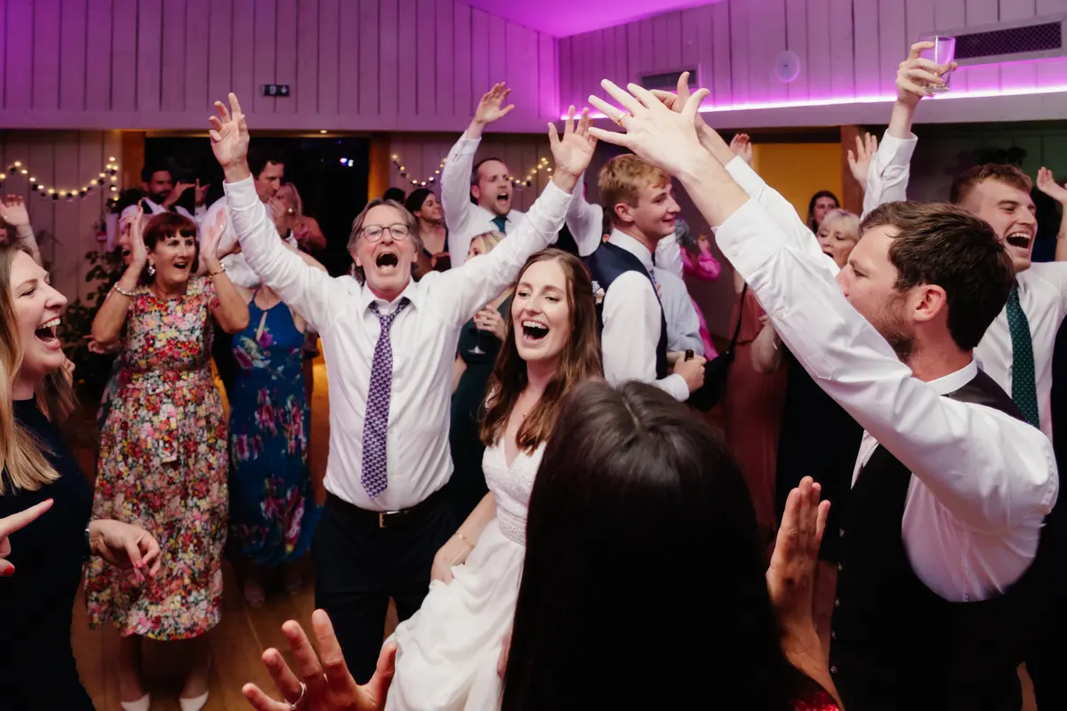 Bride and guests singing with arms in the air on a dance floor lit pink