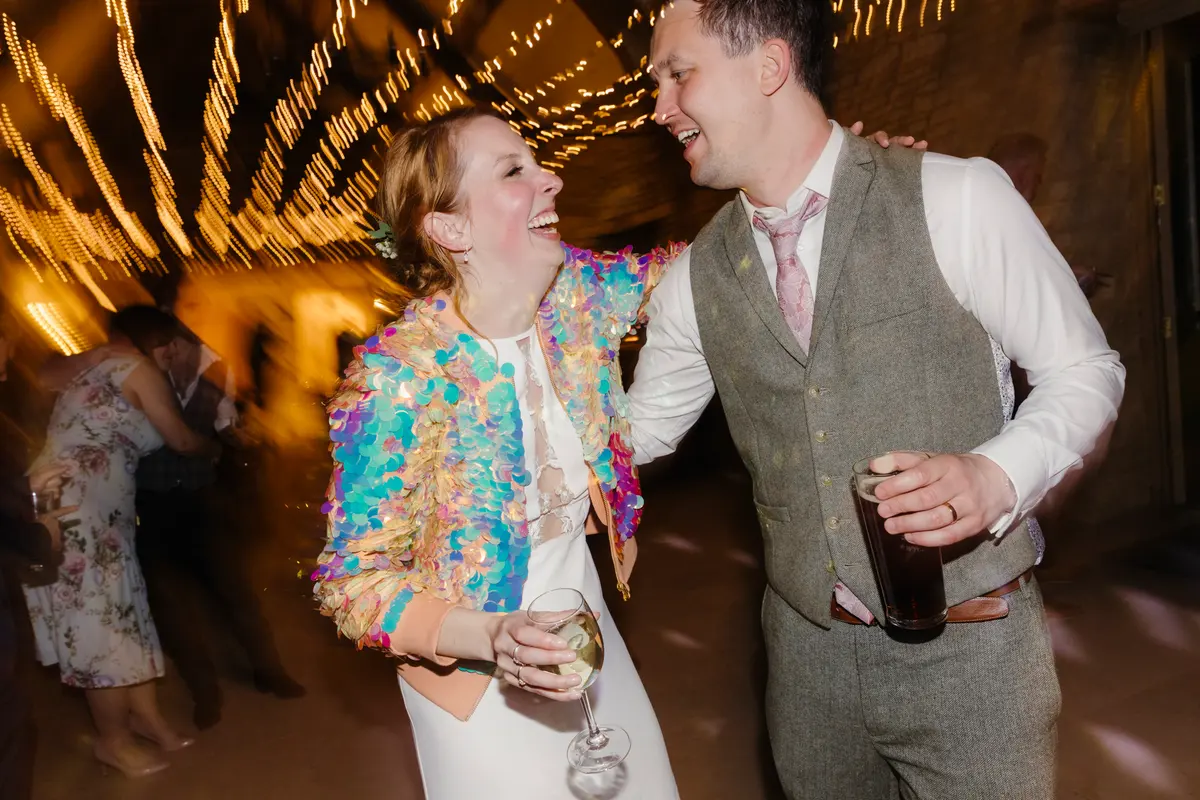 Bride in a sequinned jacket laughing and dancing with the groom at Caswell House