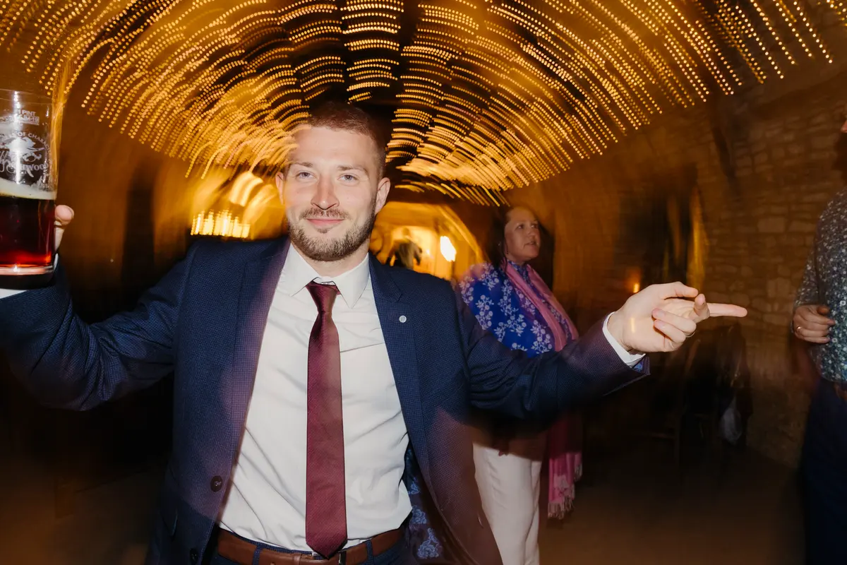Guest in a navy suit pointing and holding a pint under a tunnel of fairy lights at Caswell House