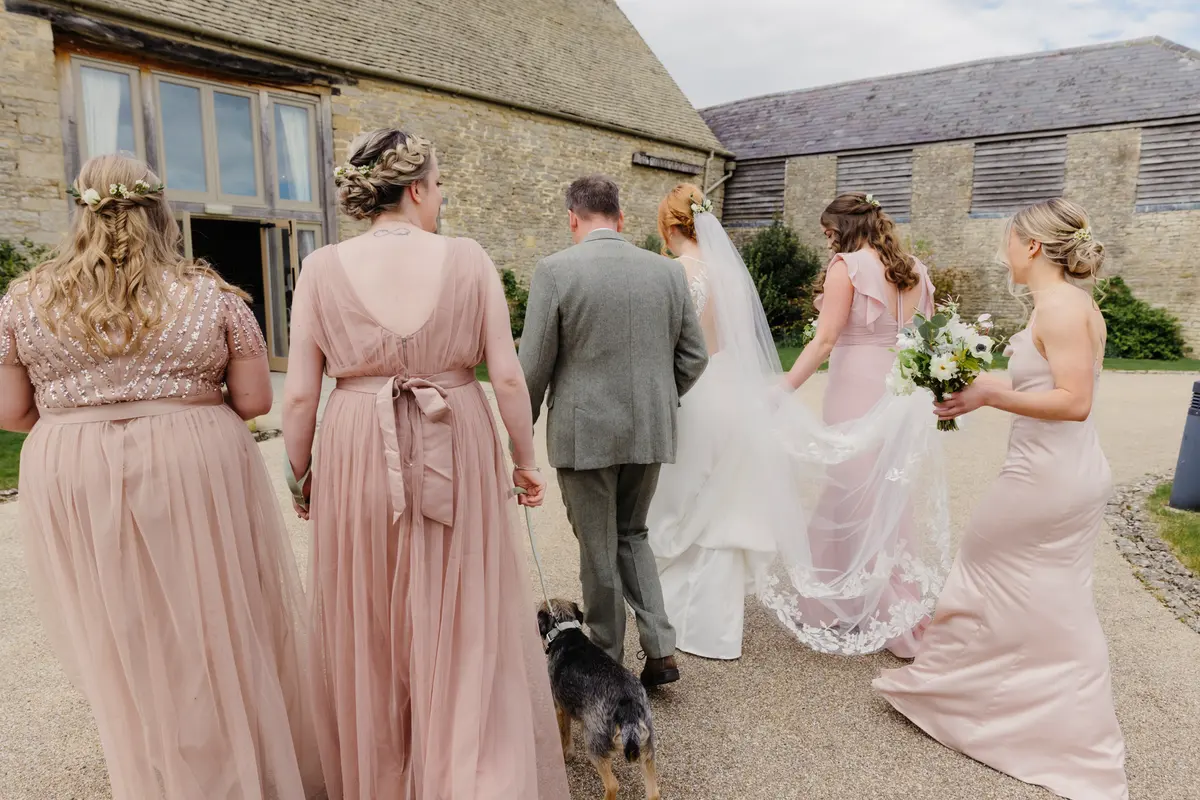 Bride walking towards Caswell House with her father, bridesmaids in blush pink and a small dog