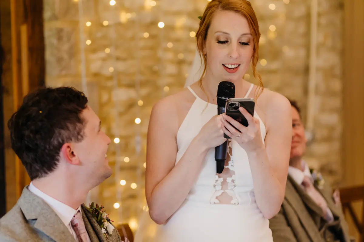 Bride giving a speech from her phone as the groom smiles up at her at Caswell House