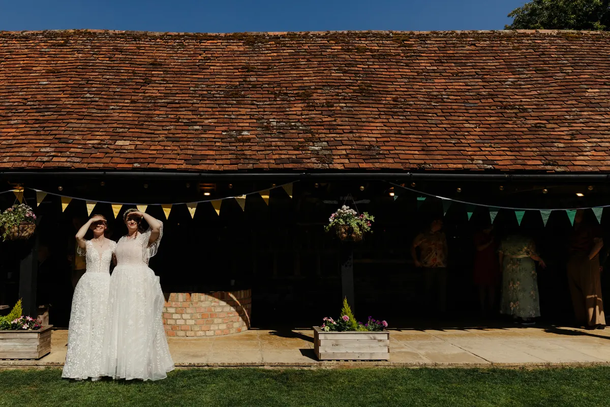 Two brides in white lace gowns shading their eyes in the sun outside the bunting-hung barn at Lains Barn