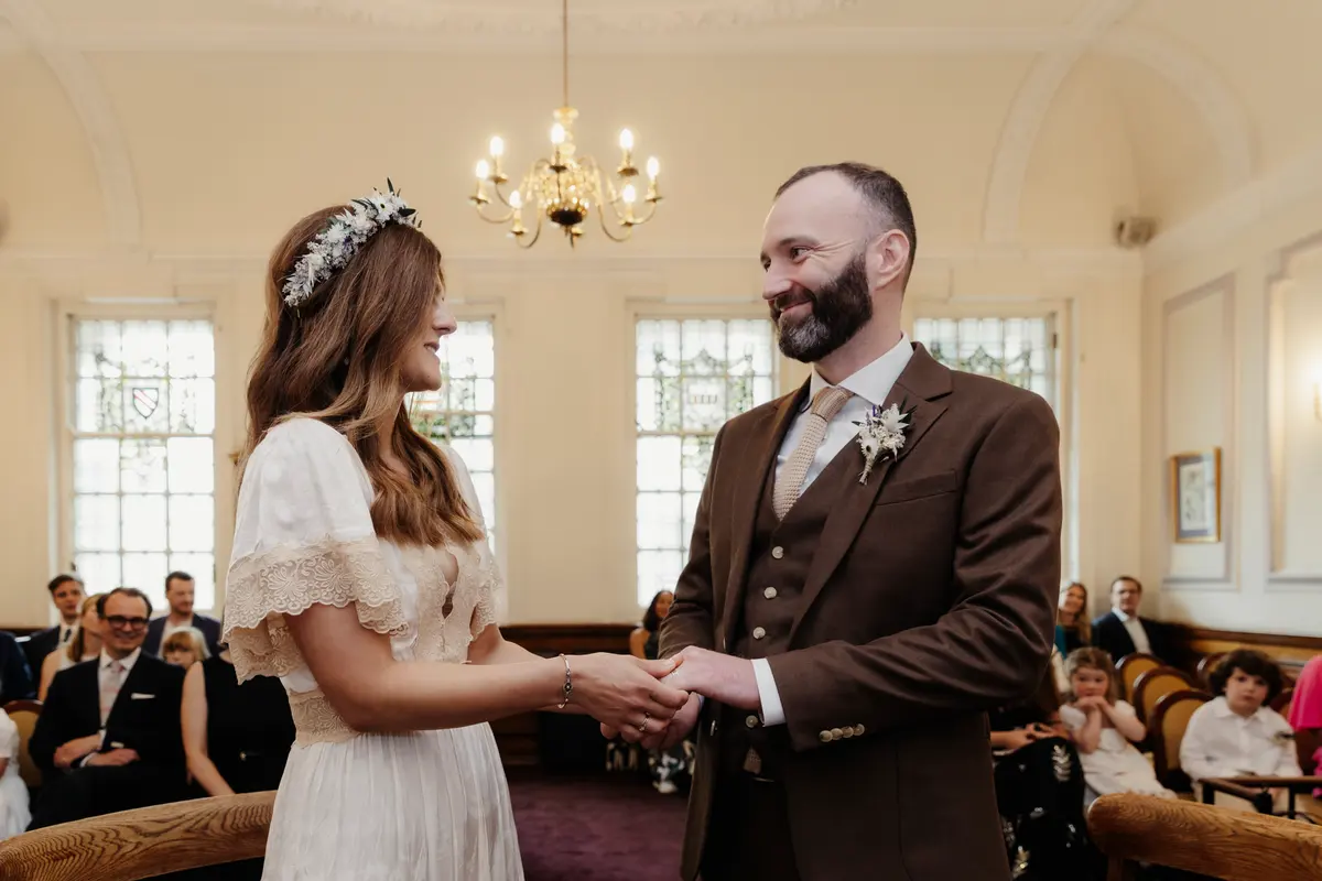 Bride and groom holding hands and smiling during their ceremony in a chandeliered room