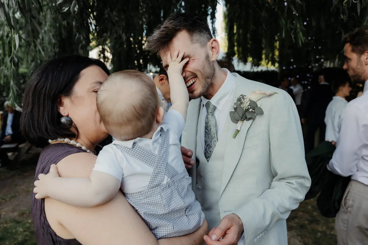 Groom laughing as a baby in a woman's arms reaches up and covers his face