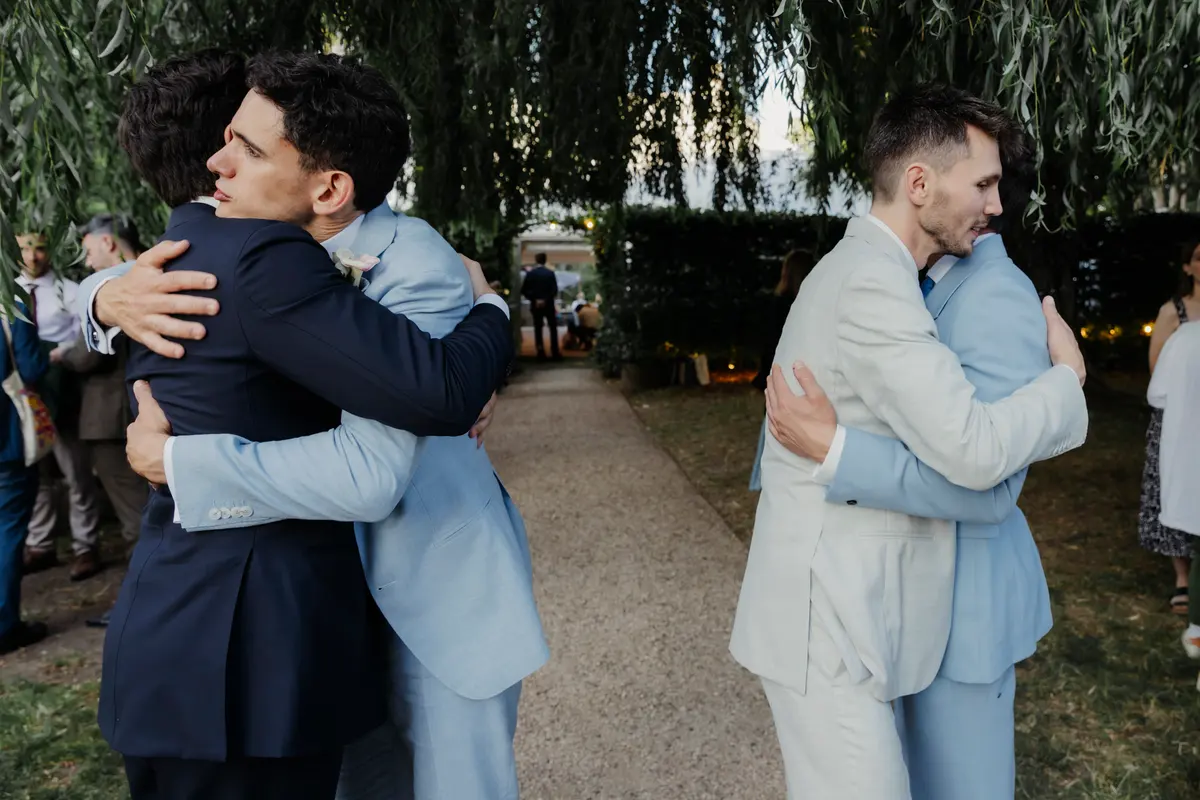 Two grooms each hugging a guest under willow trees at the Perch, Oxford