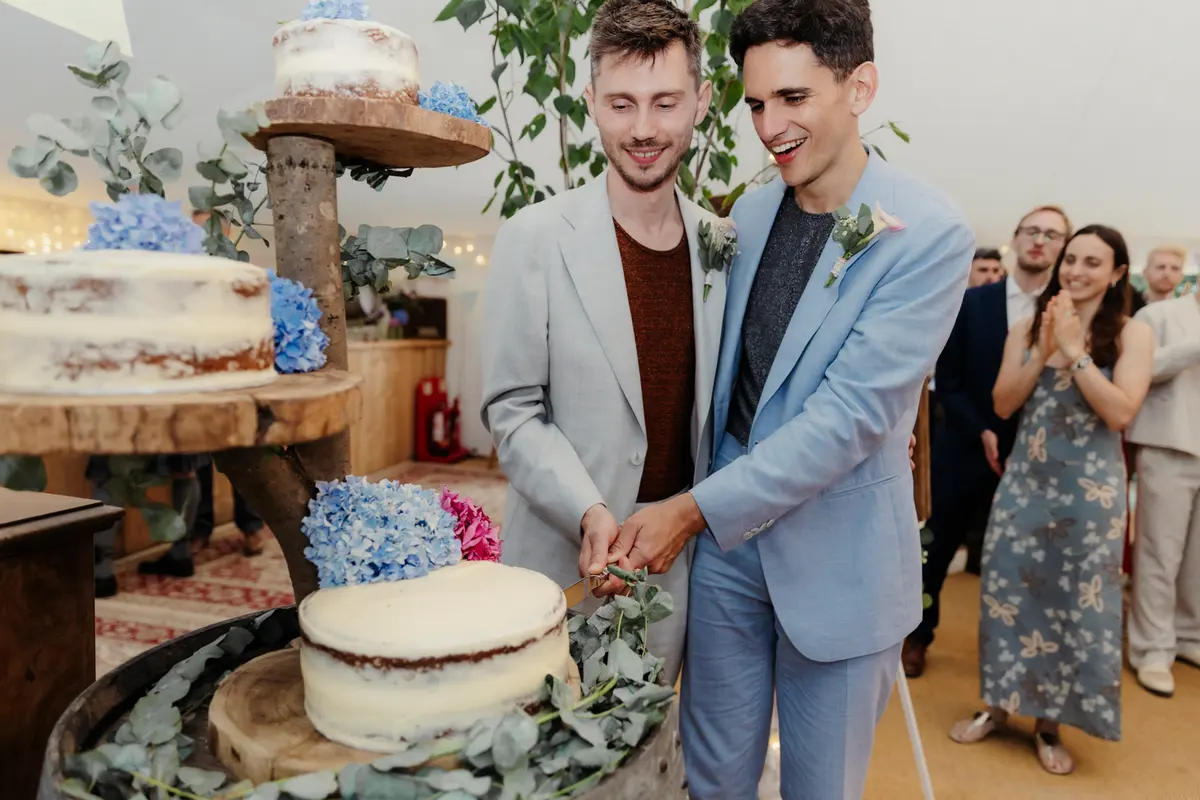 Two grooms cutting a naked cake decorated with blue hydrangeas in a marquee at the Perch, Oxford