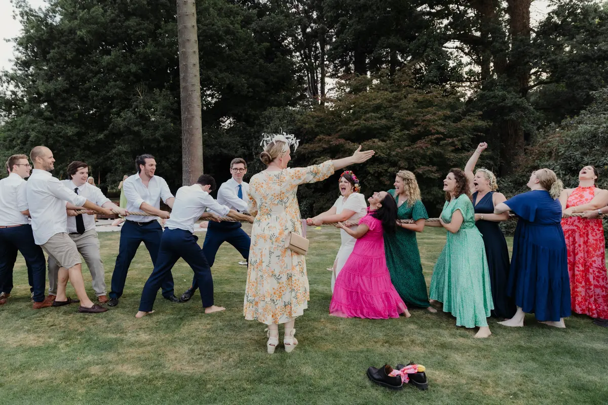 Woman in a floral dress leading a tug of war between men and women on a lawn, shoes left on the grass
