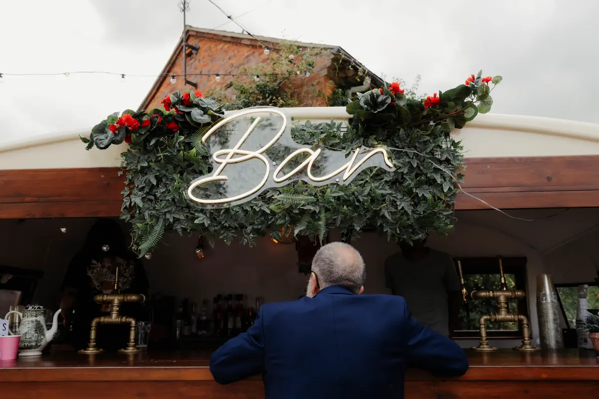 Guest leaning on an outdoor bar under a lit neon Bar sign framed with foliage and red flowers