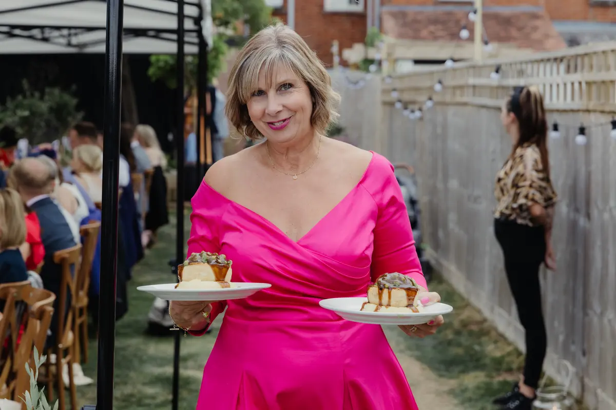 Woman in a pink dress smiling as she carries two plates of dessert past guests in a garden